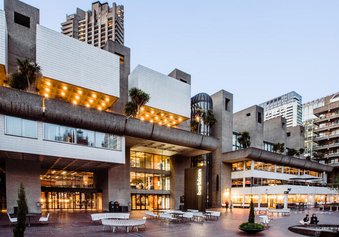 A photo of the Lakeside at the Barbican from the left hand side, at dusk with lower light and orange lights coming from the building