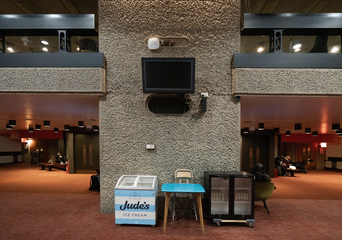 An ice cream fridge, alnog with a table and chair, sits in front of a concrete pillar in the Barbican foyer