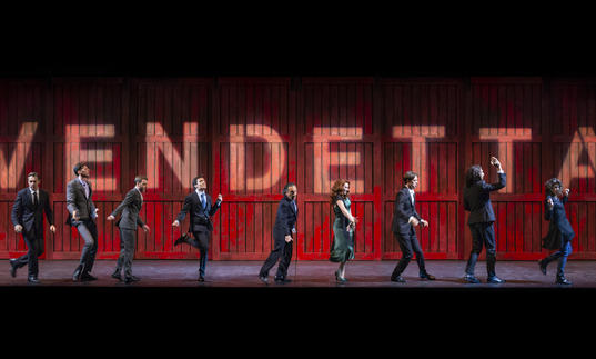 Ten people stand in various poses in front of a red wall which spells out 'vendetta' 