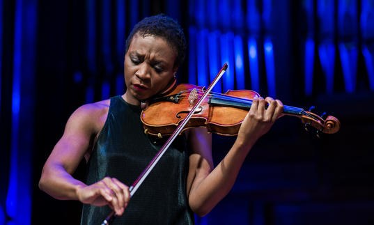 Tai Murray stands playing the violin, in front of organ pipes with a blue light on them