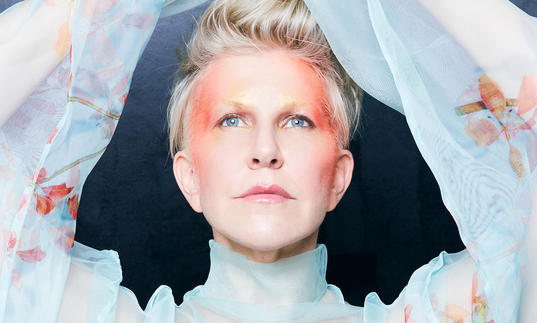 Joyce DiDonato in a blue floaty top with her arms above her head, with red face paint on her temple and the sides of her face