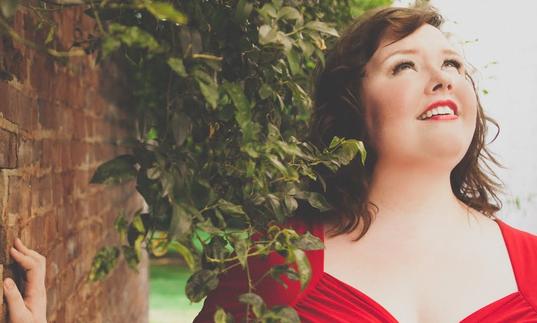 Jamie Barton in a red dress beside a brick wall and a plant draping over her shoulder