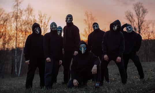 Eight people dressed in black stand in a group in front of a sunset in a field. They are wearing face masks. 