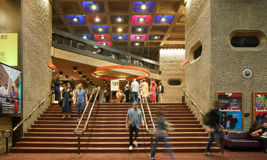 Blurred people walk down the stairs from the Barbican foyers, with bright ceiling panel lights