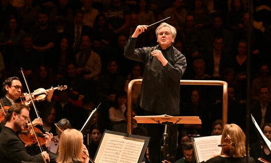 Sir Antonio Pappano conducting the LSO on the Barbican stage, holding a baton, with LSO string players in the foreground of the image and audience members in the background.