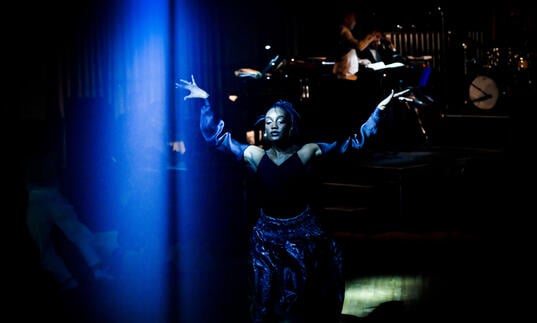 A person wearing black and blue walks with their hands in the air across the stage as a live band plays behind them.