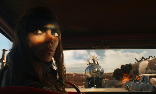 A woman in an apocalyptic landscape sits in a dark vehicle reversing. 