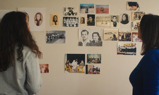 Two women look at a wall that has photographs and notes pinned on to it. 