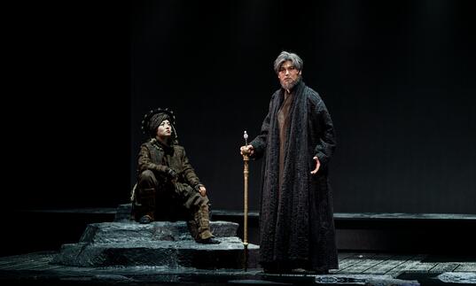 Lear stands with a wooden stick looking out at the audience while another person dressed in a dark green headdress sits on a stone beside him.