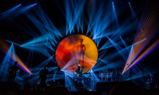 Brit Floyd performing on stage, with blue lasers and an orange sun.