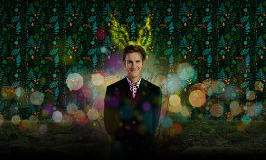 A person in a suit has a pair of glowing antlers above their head while standing against a red and green background.