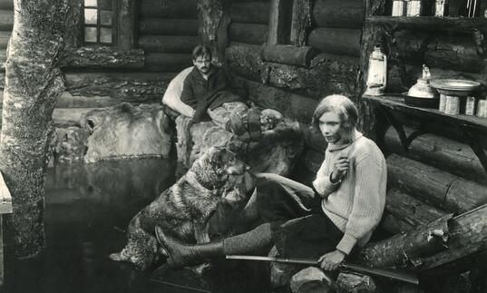 A black and white image of a man and a woman sitting by a log cabin with a large dog.