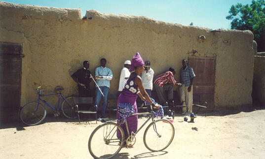 A woman passes a group of men on a bicycle in a desert village. 