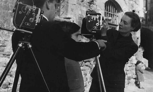 Margaret Tait stands by two large, old fashioned cameras with a man next to her in an image from the 1970s.