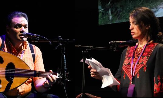 A person plays the oud on stage while another reads into a microphone from a piece of paper.