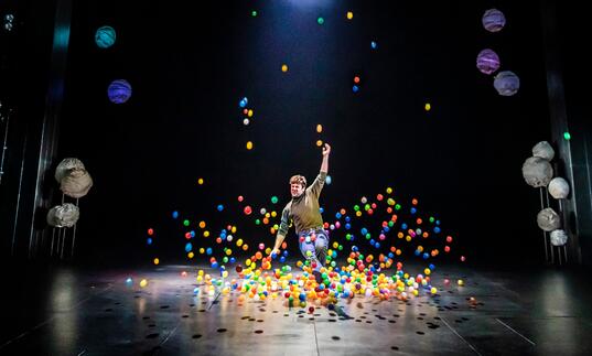 A person moves on stage around lots of multicoloured balls that bounce around them. Blue lanterns hang above them.