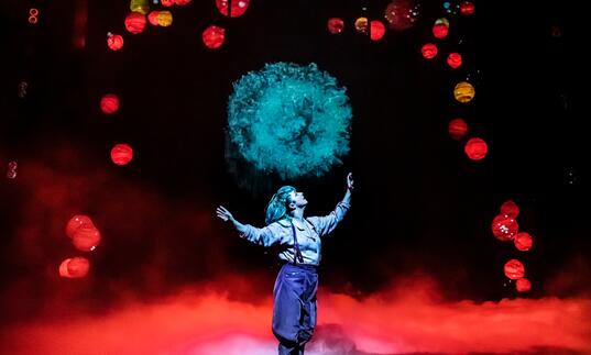 A person stands on stage looking up at a burst of aqua blue light. Red and yellow lanterns hang above her.