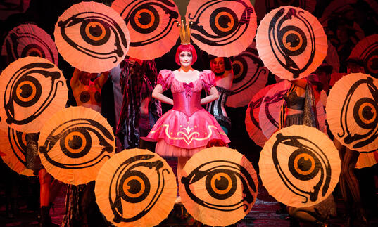 An actor stands in the middle of the stage in a pink dress, surrounded by large eyes. 