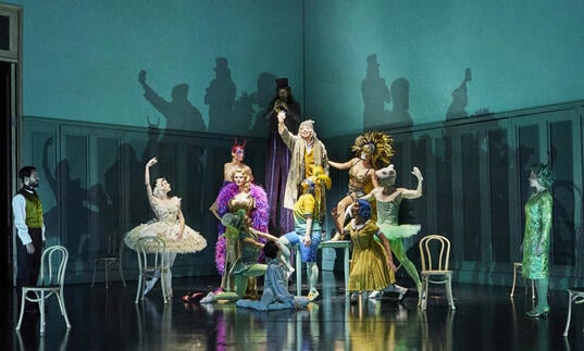 A group of ballet dancers in very colourful clothing pose on a turquoise stage.