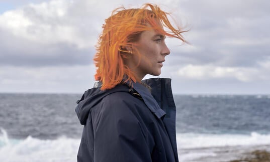 A woman with orange hair stands in front of a stormy sea. 