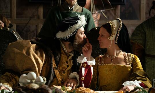 A man and a woman in tudor dress gaze into each other's eyes at a banquet, as the woman touches the man's nose.