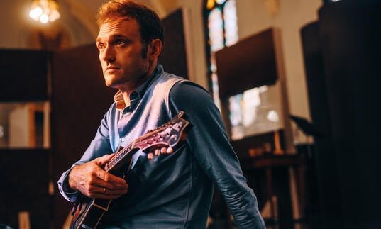 Chris Thile sitting in a room holding a mandolin