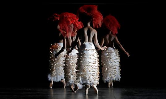 A group of dancers stand on point in a group. They wear red headdresses and white skirts that are decorated with pegs.