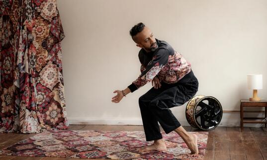 Hetain Patel holds an angular pose while performing on a red fabric in a room with a wooden floor.