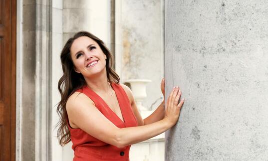 Louise Alder standing by a large concrete pillar, smiling and looking upwards