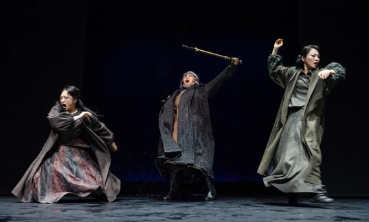 Three performers rage on stage in a production of Lear. They wear muted coloured clothing against a black background.