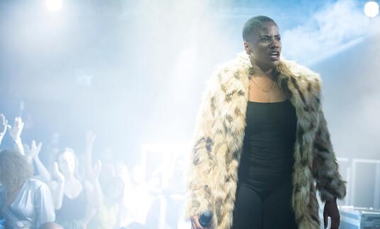 A person in a fur coat stands on stage as people dance in the background in a dark room lit by haze.