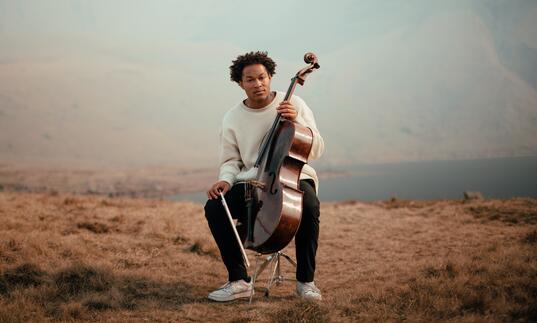 Sheku Kanneh-Mason sitting with his cello in a mountainous landscape, with a lake in the distance behind him