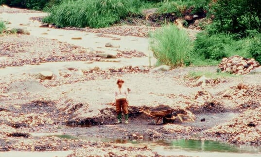 A young person sits on a rock in front of a wide, calm river.