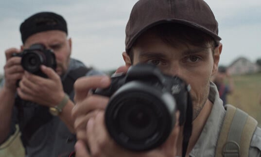 A young man with a professional camera looks ahead, whilst another man with a camera stands behind him, ready to shoot. 