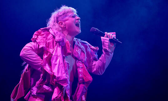 Peaches in pink stage lighting sings while holding a microphone 