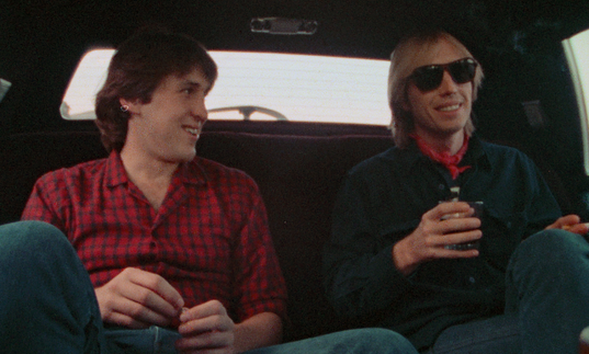 Two men sit in the back of a cab, chatting.