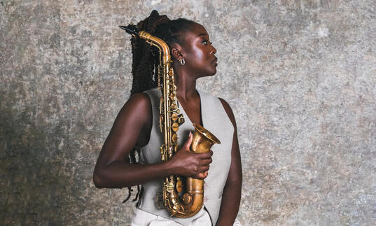 A profile shot of Cassie Kinoshi holding her saxophone in front of a concrete wall