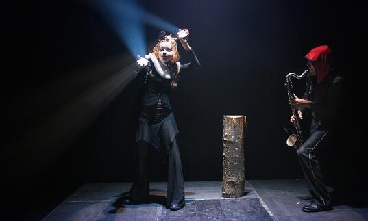 A person in a mask dances by a tree trunk while another person plays the saxophone. 