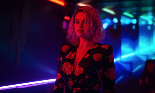 A woman stands in a blue, dark corridor, bathed in red light. 