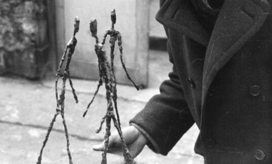 A black and white photo of Alberto Giacometti in his studio holding his sculpture, Three Men Walking.