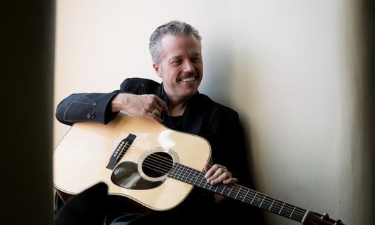 Jason Isbell smiling, sitting with guitar on lap