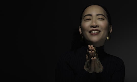 Helen Sung against black background, smiling with eyes closed and hands in prayer position