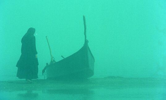 A misty scene with a bright green tint, showing a woman standing next to a moored boat.