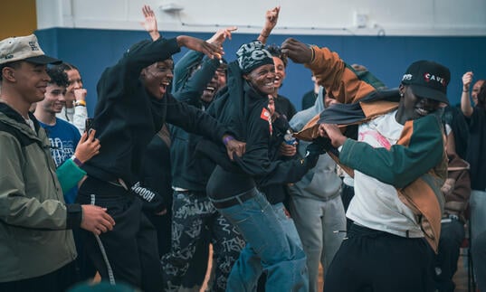 A group of hip hop dancers cheer and embrace in a rehearsal room.