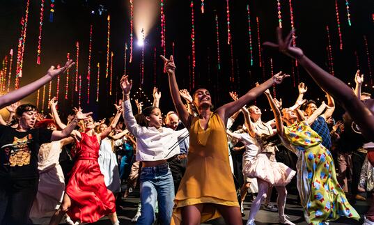 A group of dancers dressed in colourful clothing move on stage with their arms in the air.