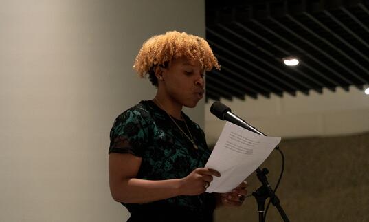 A Barbican Young Poet holds a piece of paper and reads into a microphone. 