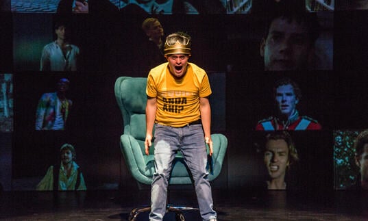 A person in a yellow shirt and blue jeans standing over a green sofa chair, shouting. They are against a background of screens with each screen showing pictures of different men.