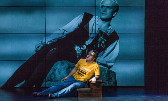 A person in a yellow shirt and blue jeans laying down on their side on a pair of stairs. They are against a background of screen which is showing a picture of another person dressed in historical clothing, also laying on their side over a rock.  