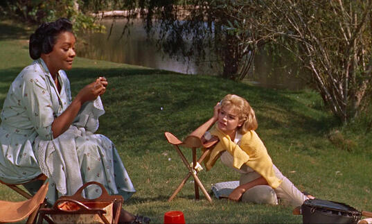 An older woman sits on the grass with a younger woman at her feet.