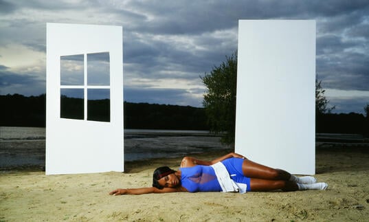 A person lies down on the beach in a blue dress. Two white walls stand behind them, one with a window. The sea can be seen in the background along with the sky.